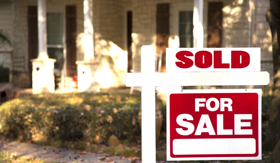 Home for sale with red and white real estate sign during the fall season.  Fall season with leaves on ground.  Front porch and windows in background.  Residential neighborhood.  Moving house, relocation concept.